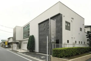 介護施設の建物外観。白と黒のモダンな3階建て構造で、緑の植栽が配置されている。